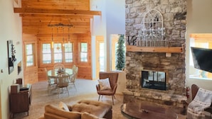 A spacious living room with a stone fireplace, wooden ceiling beams, and a large window overlooking a deck.