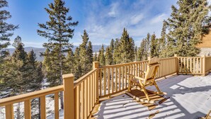 Une terrasse en bois dont le plancher est recouvert de neige, entourée de grands pins et surplombée d’un ciel bleu clair.