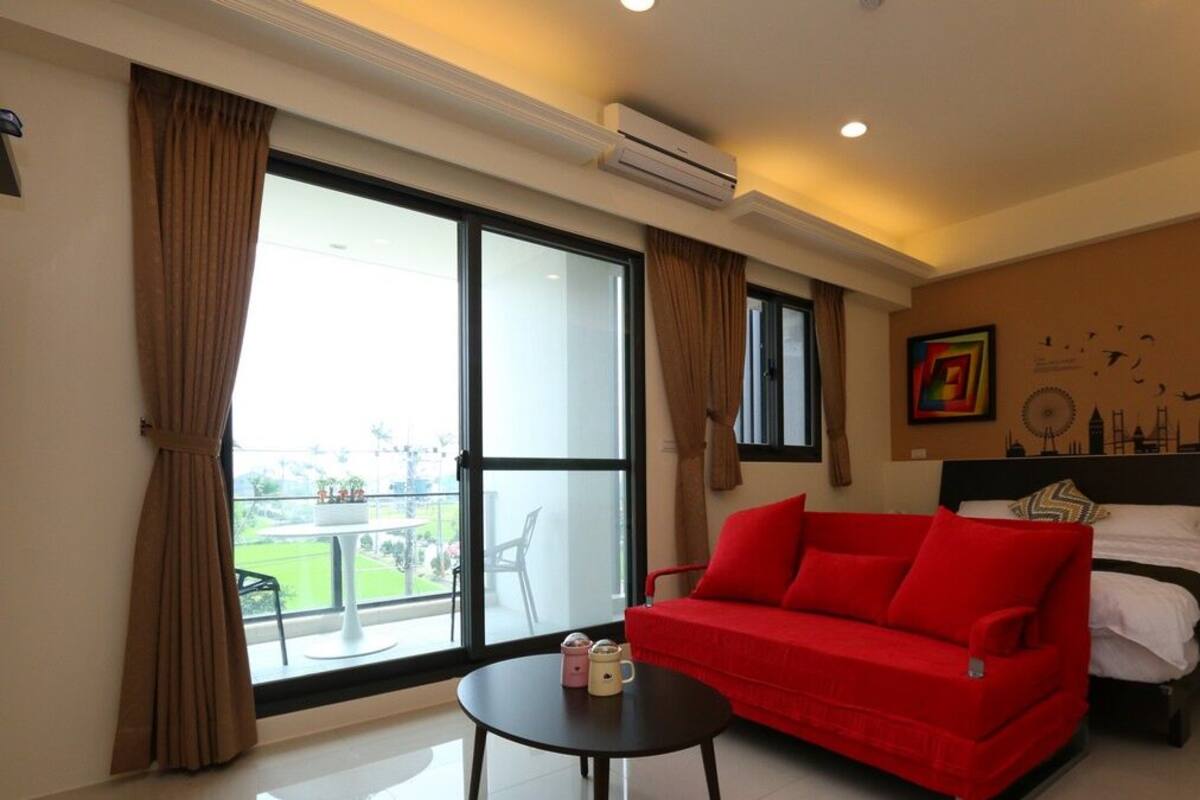 A hotel room with a bed, a red sofa, a small table with a teapot, and a view of a cityscape through large windows.