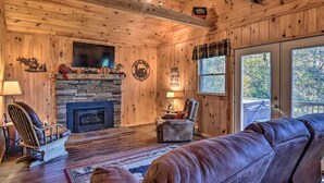 A cozy cabin interior with a stone fireplace, wooden walls, a comfortable seating area, and a large window offering a view of the outdoors. - 'Nature's Retreat' w/ Hot Tub: 7 Mi to Bryson City (Whittier)