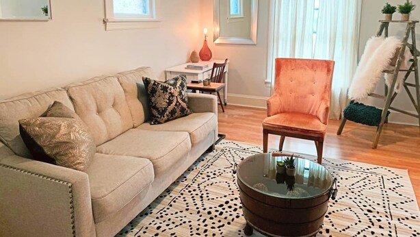 Ein Wohnzimmer mit einem beigen Sofa, einem runden Couchtisch und einem Holzstuhl. Zwei Fenster mit Vorhängen, ein Deckenventilator und ein kleiner Tisch mit einer Lampe sind ebenfalls vorhanden.