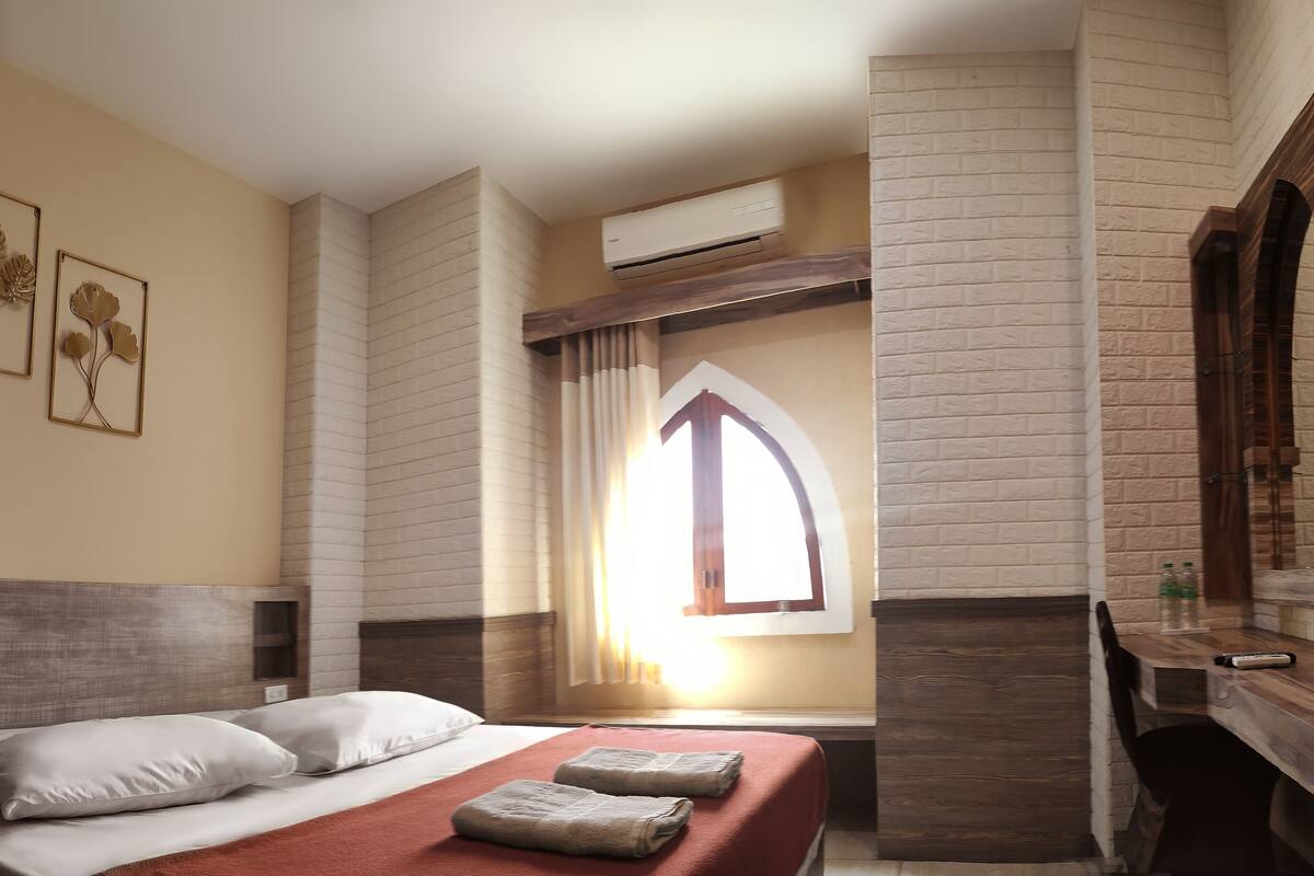 A hotel room with a bed, a wooden desk, an air conditioning unit, and a window with curtains.