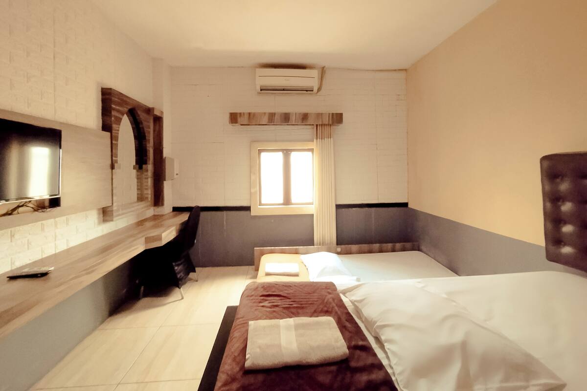 A hotel room with a bed, a TV mounted on the wall, a window, and an air conditioning unit.