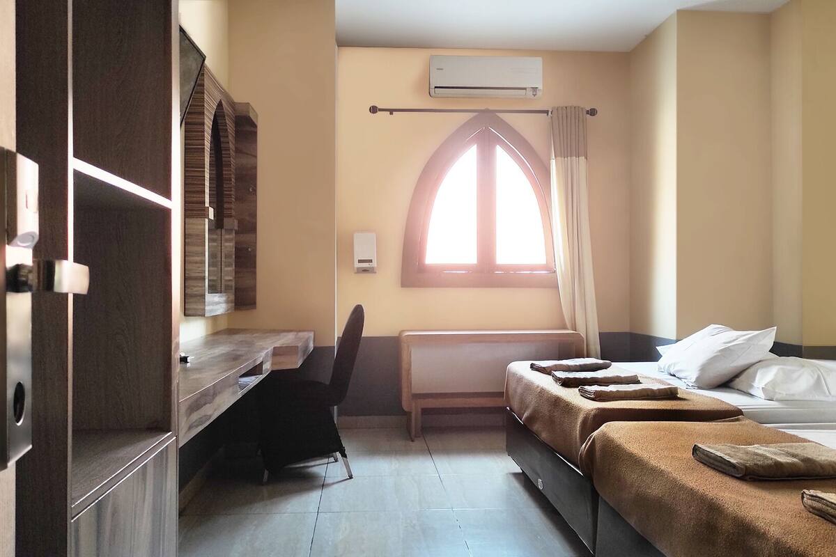 A hotel room with a desk, two beds, a window with curtains, an air conditioning unit, and a radiator.