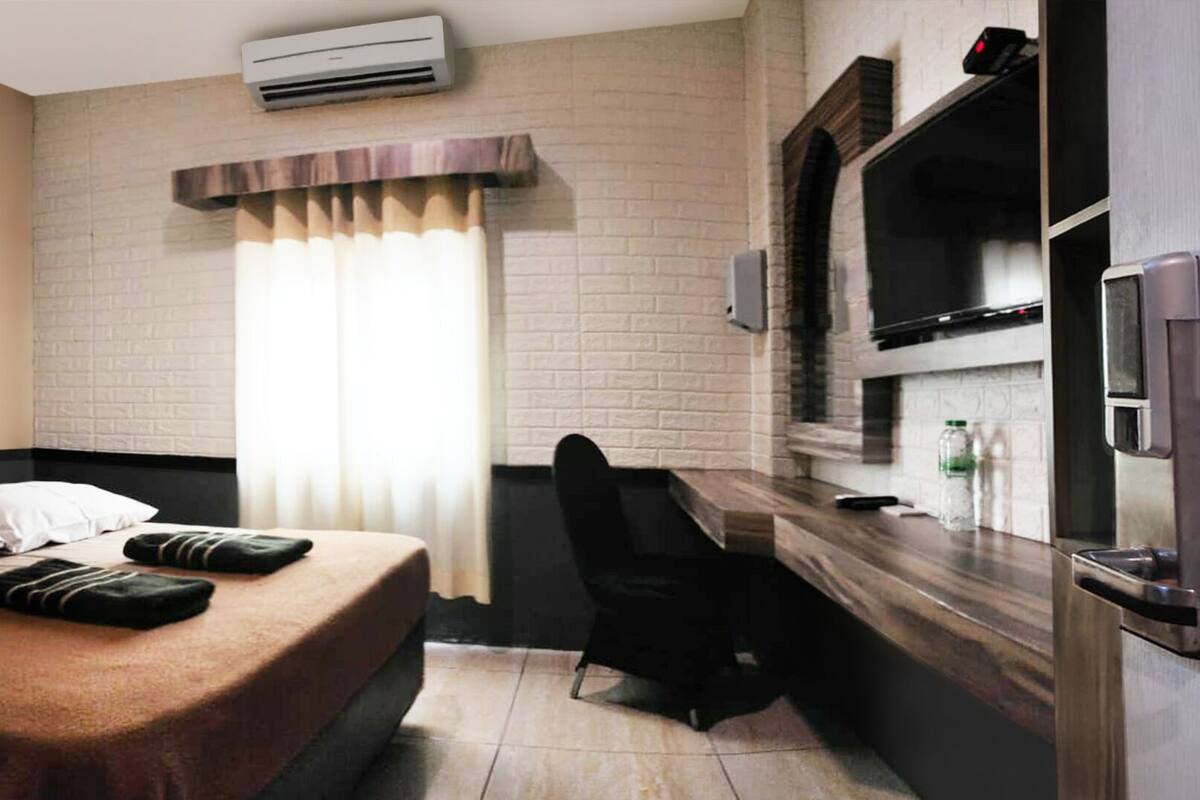 A hotel room with a bed, a desk, a chair, an air conditioner, a TV, and a window with curtains.