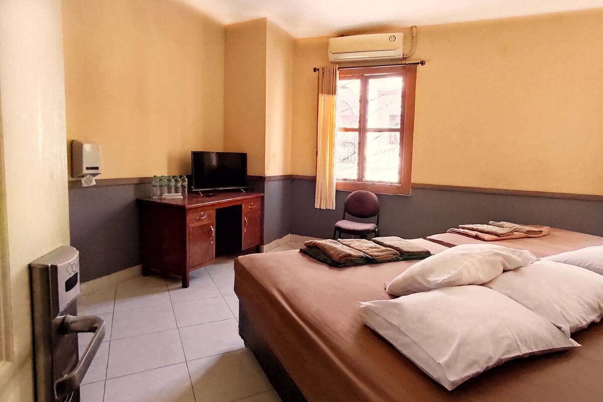 A hotel room with a bed, a desk with a television, a chair, and an air conditioning unit.