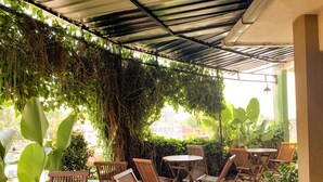 An outdoor patio with wooden chairs and tables, surrounded by lush greenery and a metal roof.