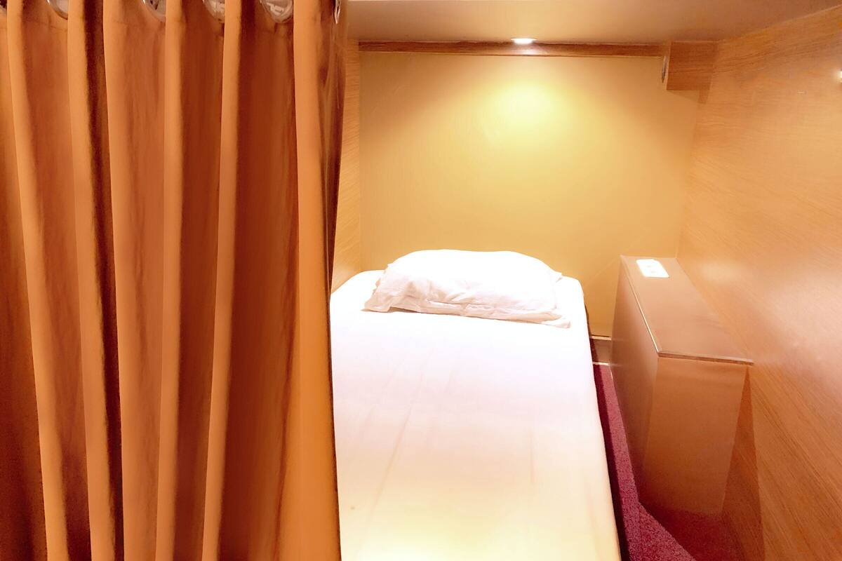 A small, compact room with a single bed, a wooden nightstand, and a curtain.