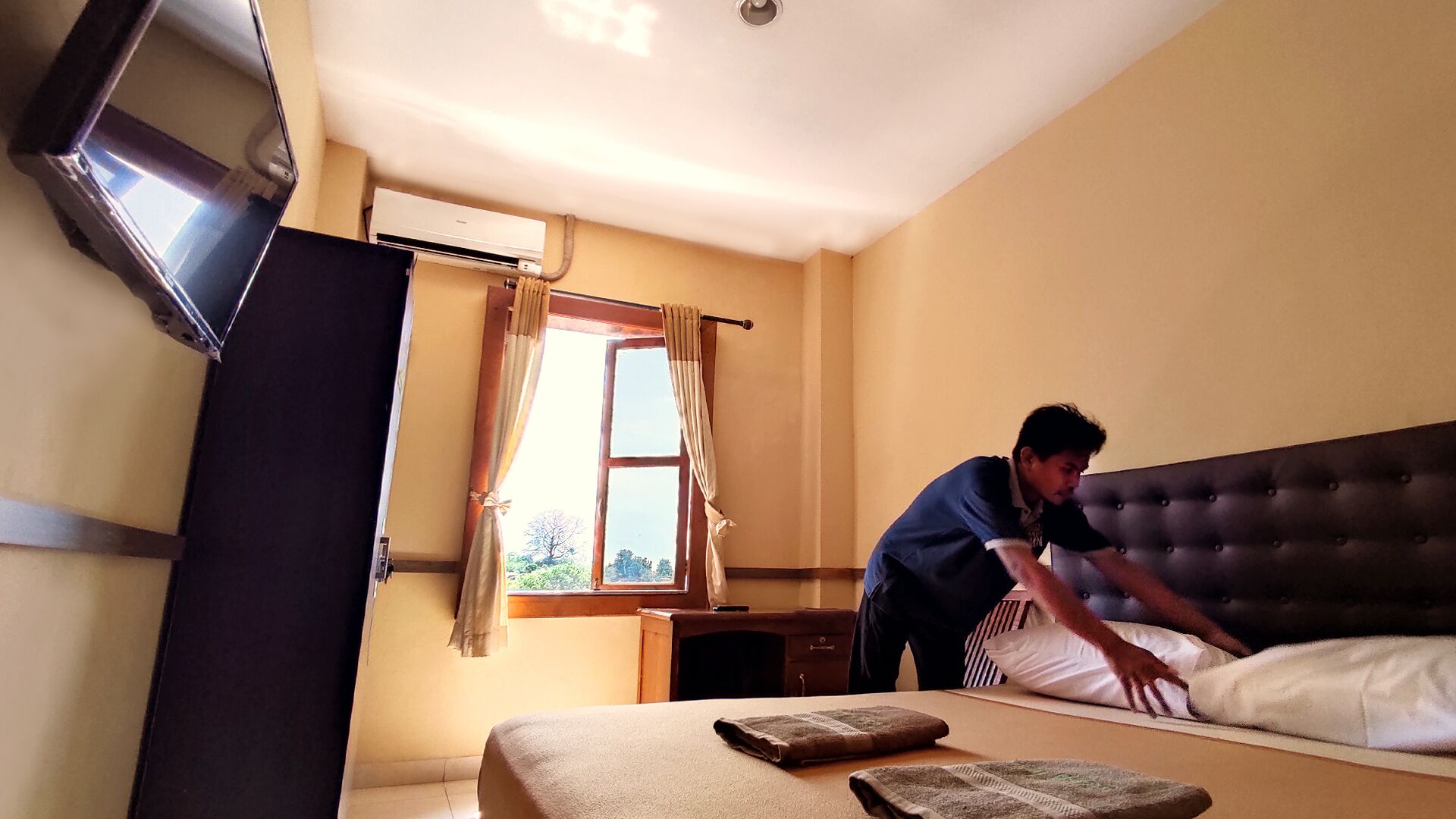 Een hotelkamer met een bed, nachtkastje, raam met gordijnen en een wandairconditioning.