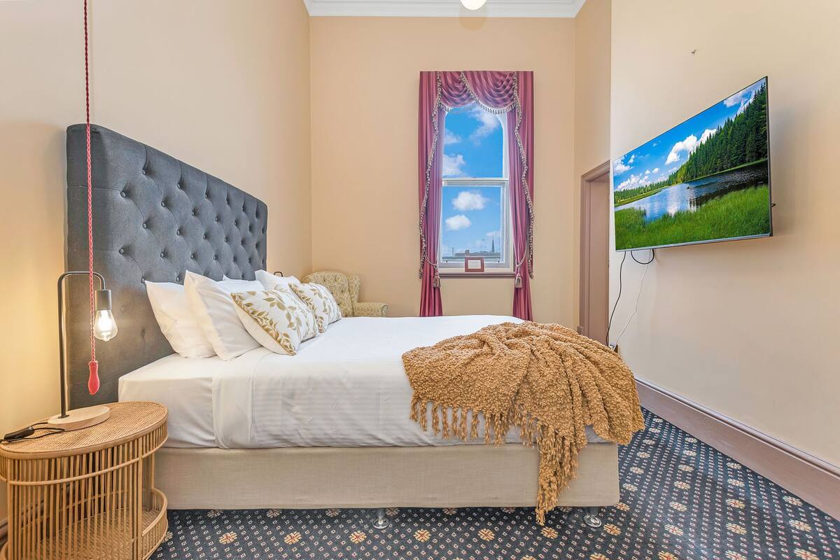 A bedroom with a large bed, a TV mounted on the wall, a window with curtains, and a patterned floor.