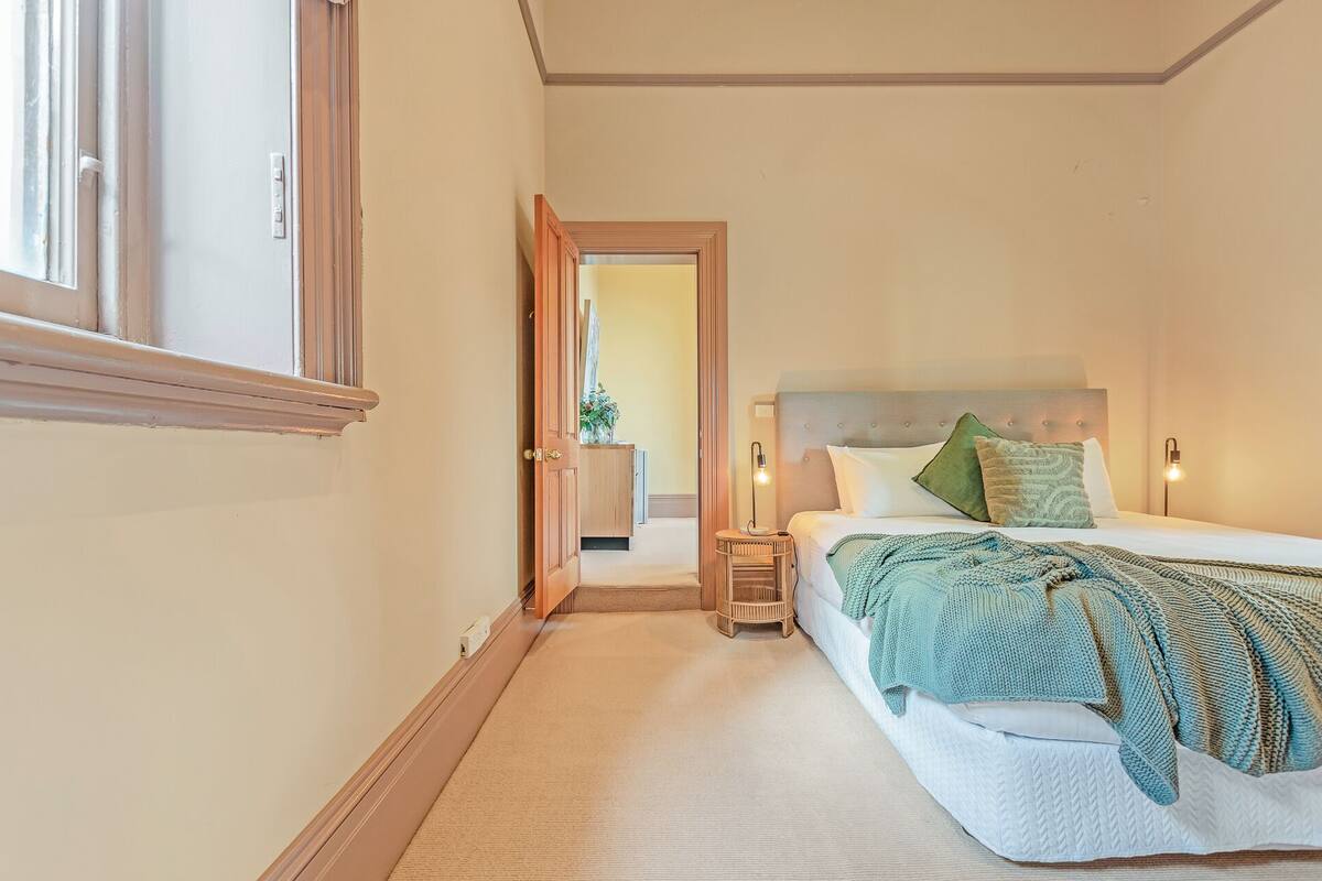 A bedroom with a bed, bedside lamps, a nightstand, and a door leading to another room.