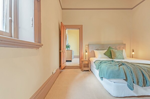 A bedroom with a bed, bedside lamps, a nightstand, and a door leading to another room. - Clocktower Suites (Echuca)