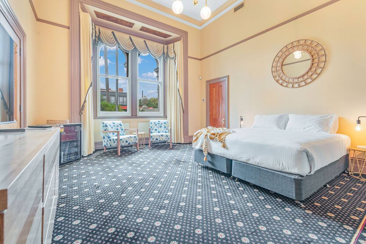 A hotel room with a large bed, a desk, and a chair. There is a window with curtains and a view of the outside.