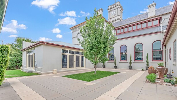 A spacious courtyard with a well-maintained lawn, a large tree, and a building with arched windows and a clock tower. - Clocktower Suites (Echuca)