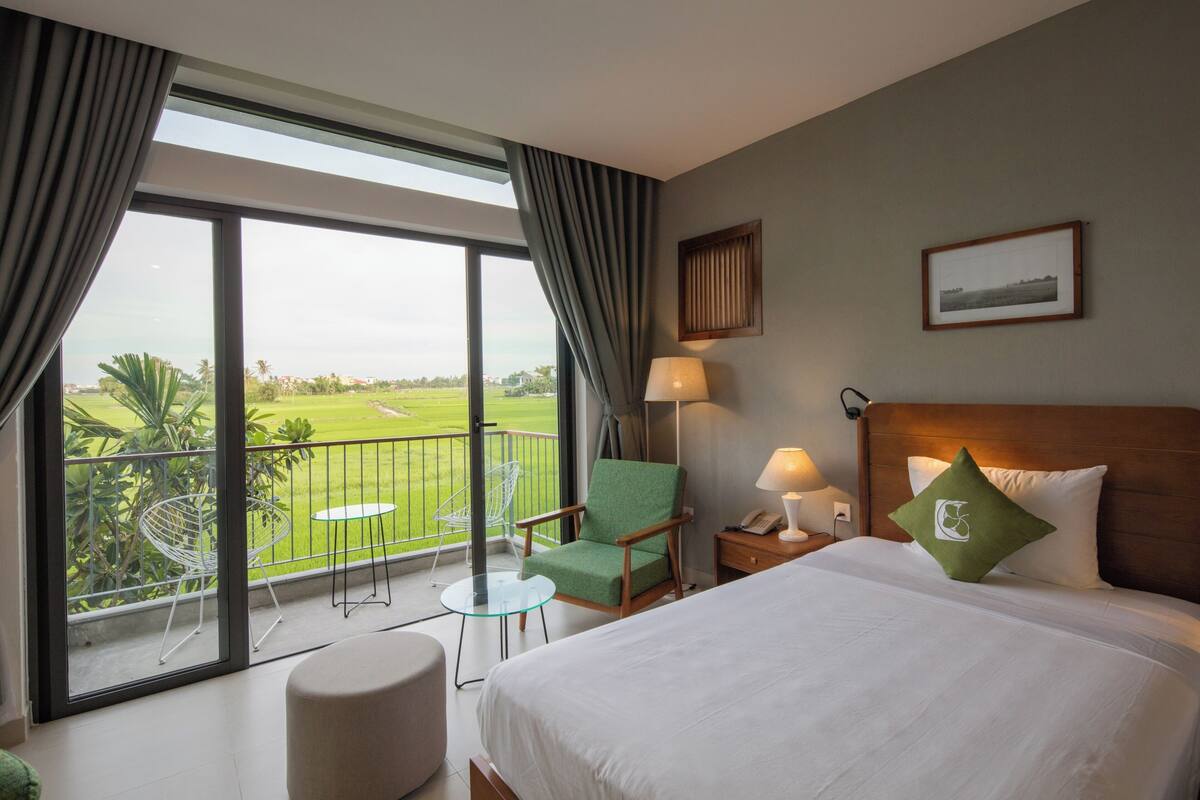 Ein Hotelzimmer mit einem großen Bett, einem grünen Sessel, einem kleinen Tisch, einem Nachttisch mit Lampe und einem Balkon mit Blick auf Grünflächen und eine weite Landschaft.