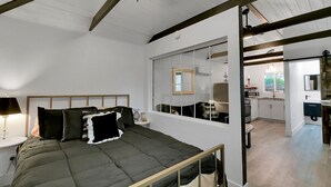 A bedroom with a bed, bedside table, lamp, and a view of the kitchen through a window.