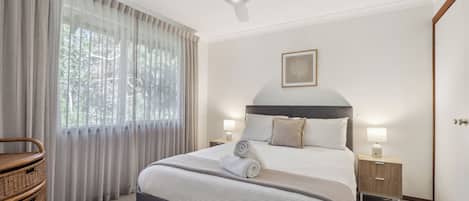A neatly arranged bedroom with a bed, bedside tables, a ceiling fan, and a window with curtains.