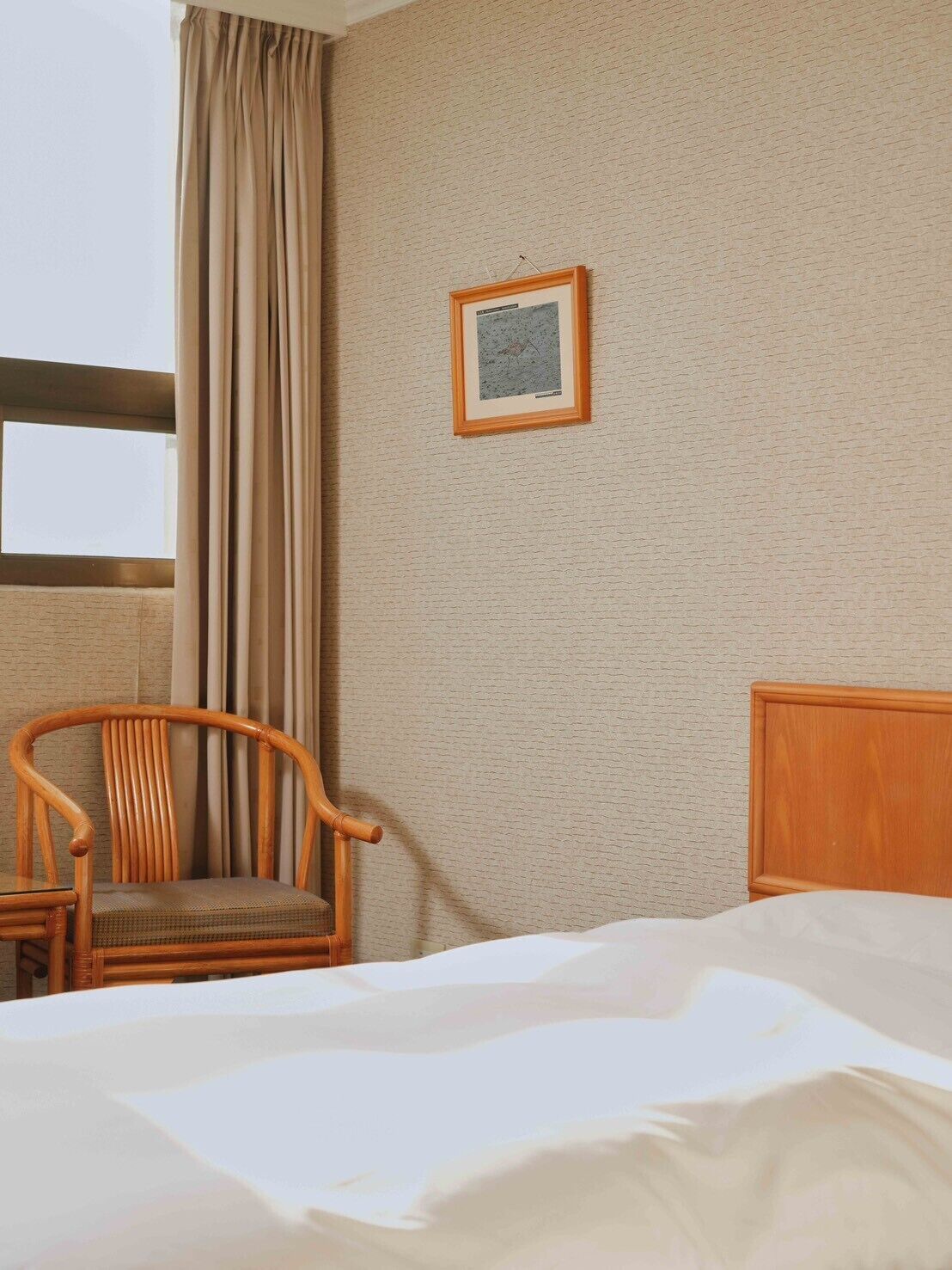 A hotel room with a bed, a wooden chair, a framed picture on the wall, and a window with curtains.
