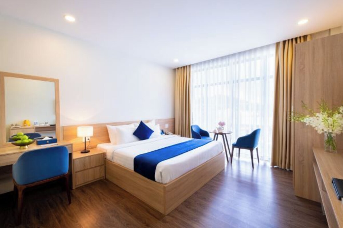 A hotel room with a large bed, a desk with a chair, a small table with a chair, and a large window with curtains.