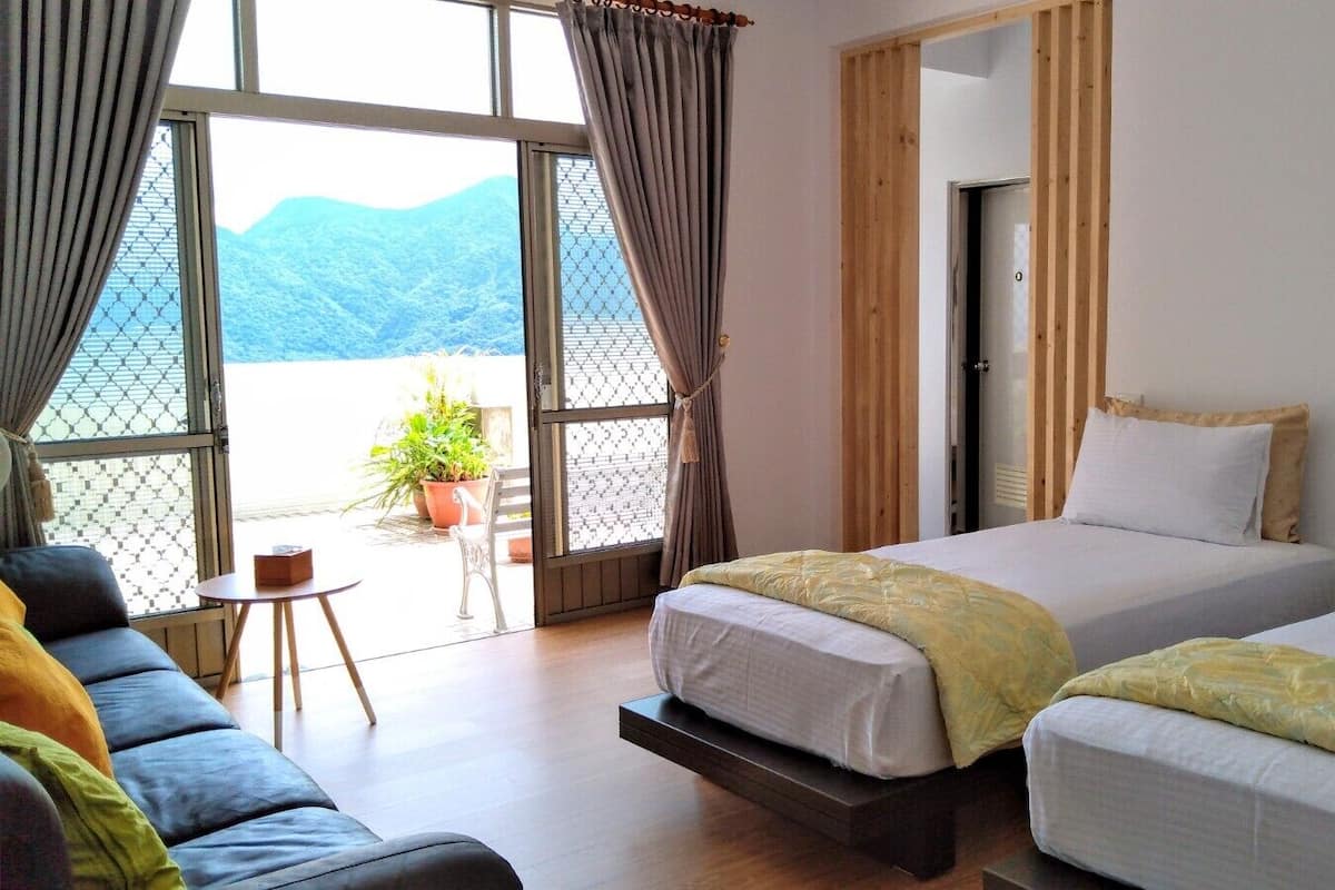 A hotel room with two beds, a sofa, and a small table. There is a balcony with a view of mountains.