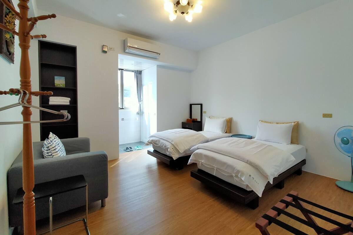 A hotel room with two beds, a grey sofa, a wooden chair, a ceiling fan, and an air conditioning unit.