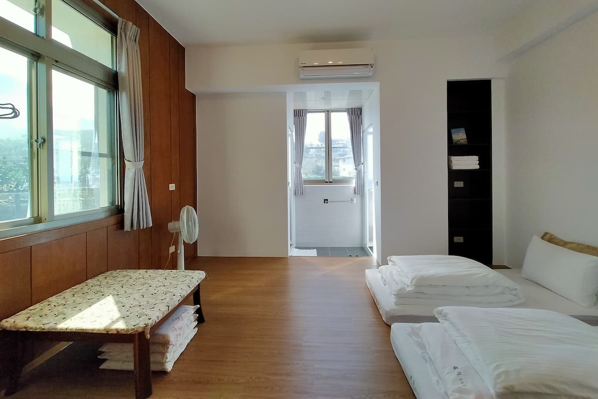A hotel room with a wooden bench, two beds, a fan, and a view through a window with curtains.