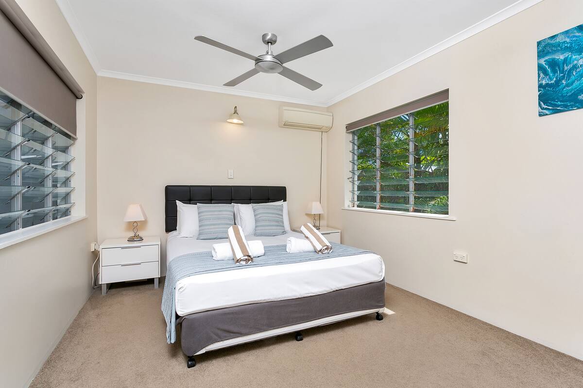 A bedroom with a bed, a ceiling fan, a window with blinds, a nightstand, and a wall-mounted air conditioner.