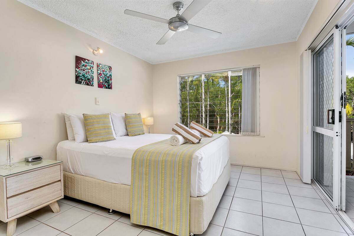 A bedroom with a bed, nightstand, lamp, ceiling fan, and a large window with a view of trees.