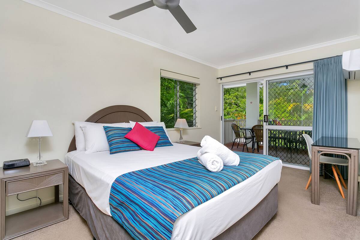 A hotel room with a bed, a desk, a chair, a nightstand, a lamp, and a ceiling fan.