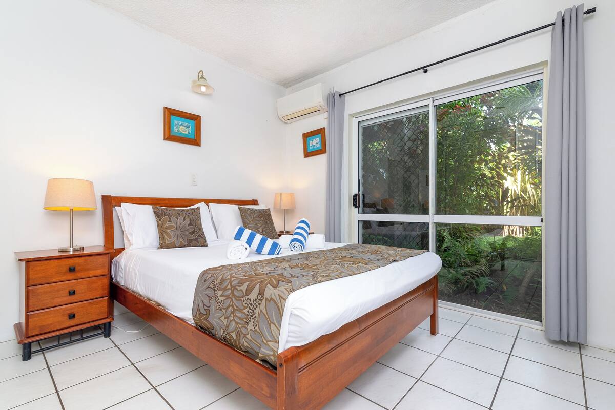 A bedroom with a wooden bed, two bedside tables, a window with a view of greenery, and a ceiling fan.