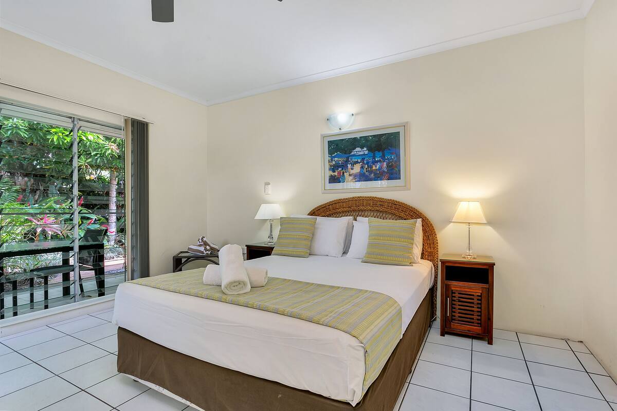 A hotel room with a bed, two bedside tables, a window with a view of greenery, and a painting on the wall.
