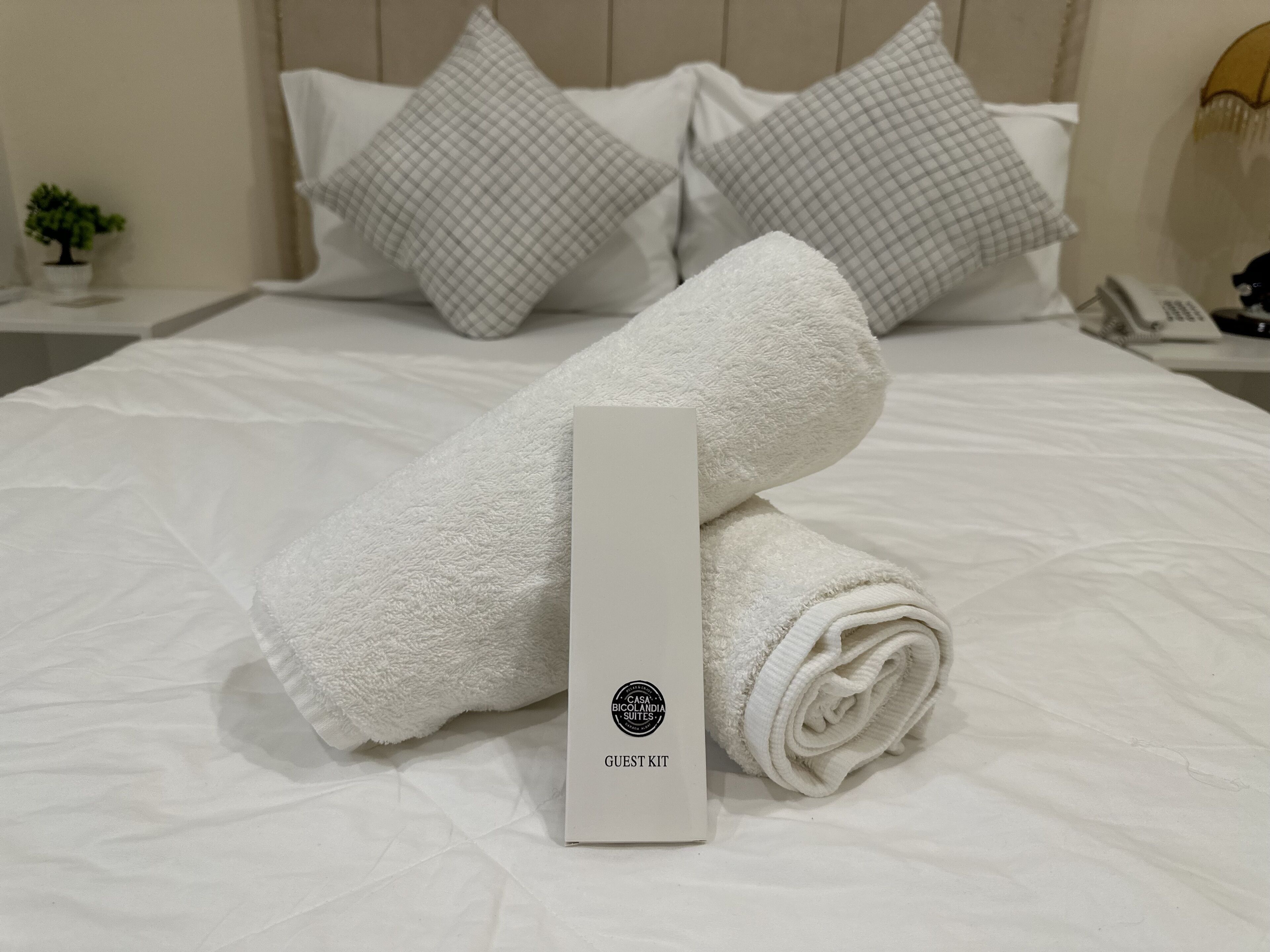 A neatly made bed with white linens, a rolled towel, and a guest kit box.