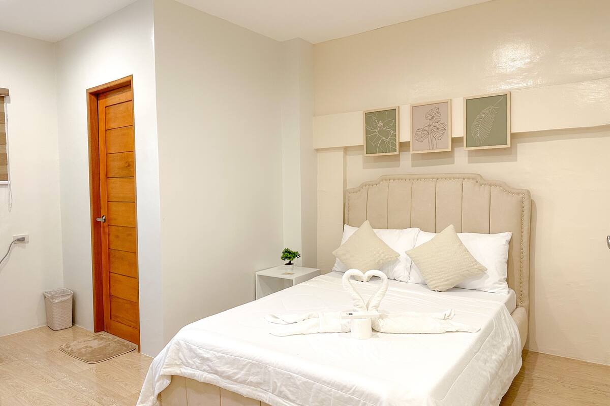A neatly made bed with white bedding and pillows, a wooden door, a small bedside table with a plant, and three framed leaf artworks on the wall.