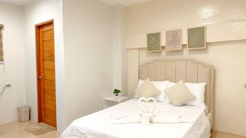 A neatly made bed with white bedding and pillows, a wooden door, a small bedside table with a plant, and three framed leaf artworks on the wall.