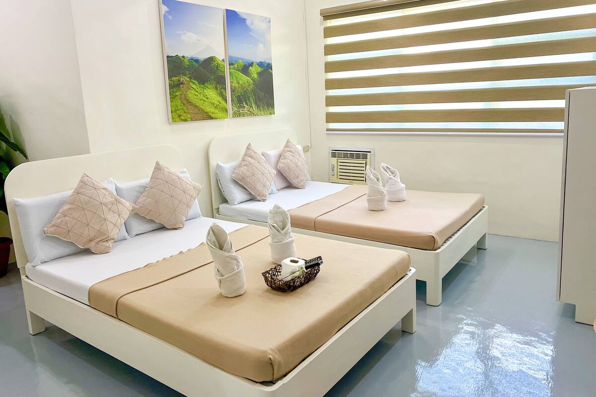 A hotel room with two beds, beige bedding, pillows, a small table with towels, a basket, and a picture of a landscape.