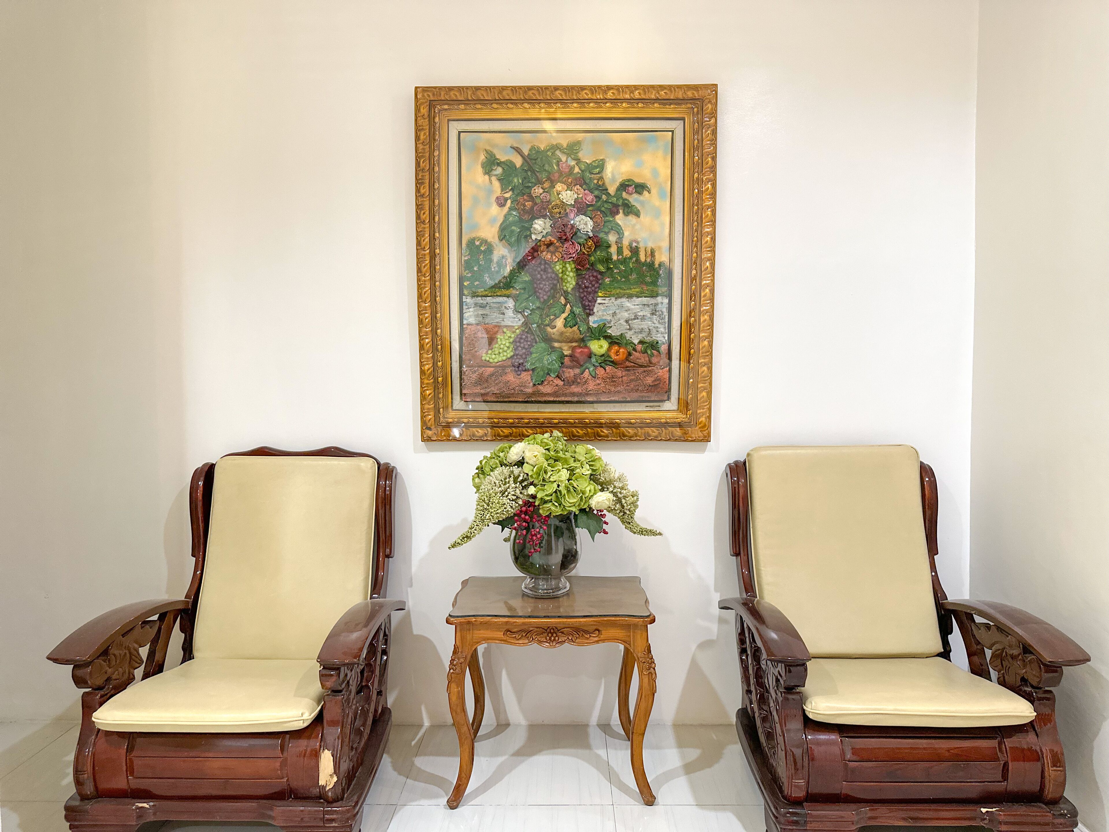 Two wooden armchairs with cream cushions, a small table with a vase of flowers, and a framed painting of a fruit tree on the wall.