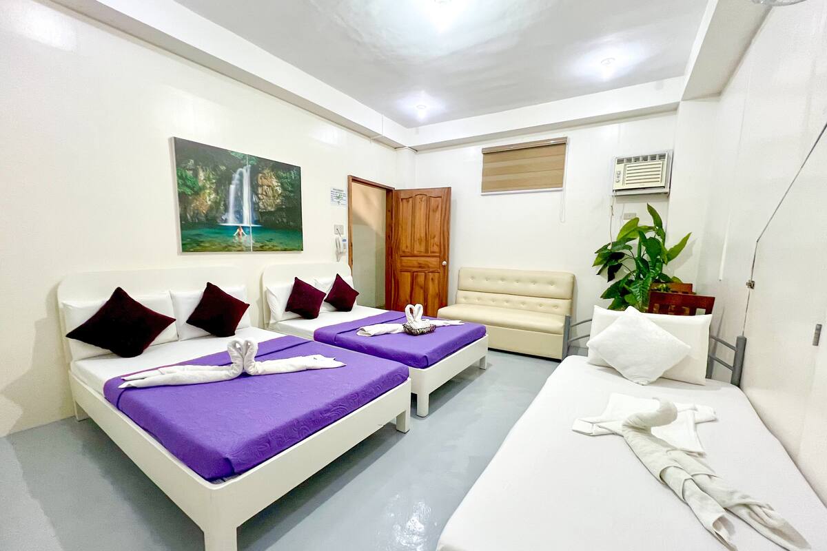 A hotel room with two beds, a sofa, a painting, a plant, and a door.