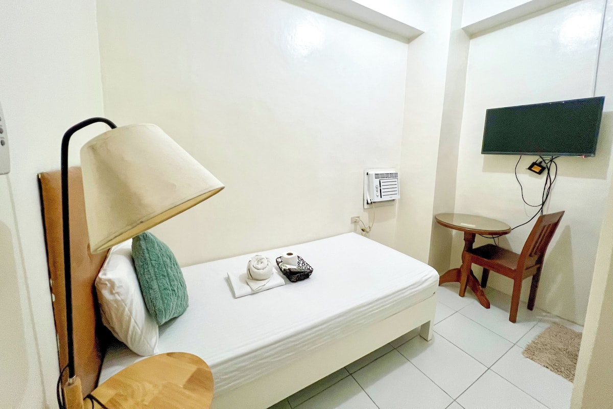 A small, clean room with a single bed, a desk, a chair, a lamp, and a TV mounted on the wall.
