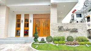 A modern building with a large wooden door, a stone wall with the name 'Casa Bicolandia Suites', and a well-maintained garden with various shrubs and a small tree. - Casa Bicolandia Suites (Daraga)