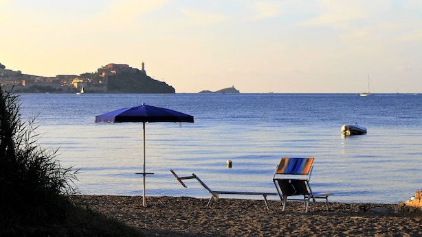 A beach with a chair and umbrella, calm sea, and a distant island with buildings and a lighthouse.