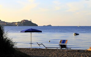 A beach with a chair and umbrella, calm sea, and a distant island with buildings and a lighthouse. - Casa Anselmi - Apartment in the countryside of Portoferraio (Portoferraio)