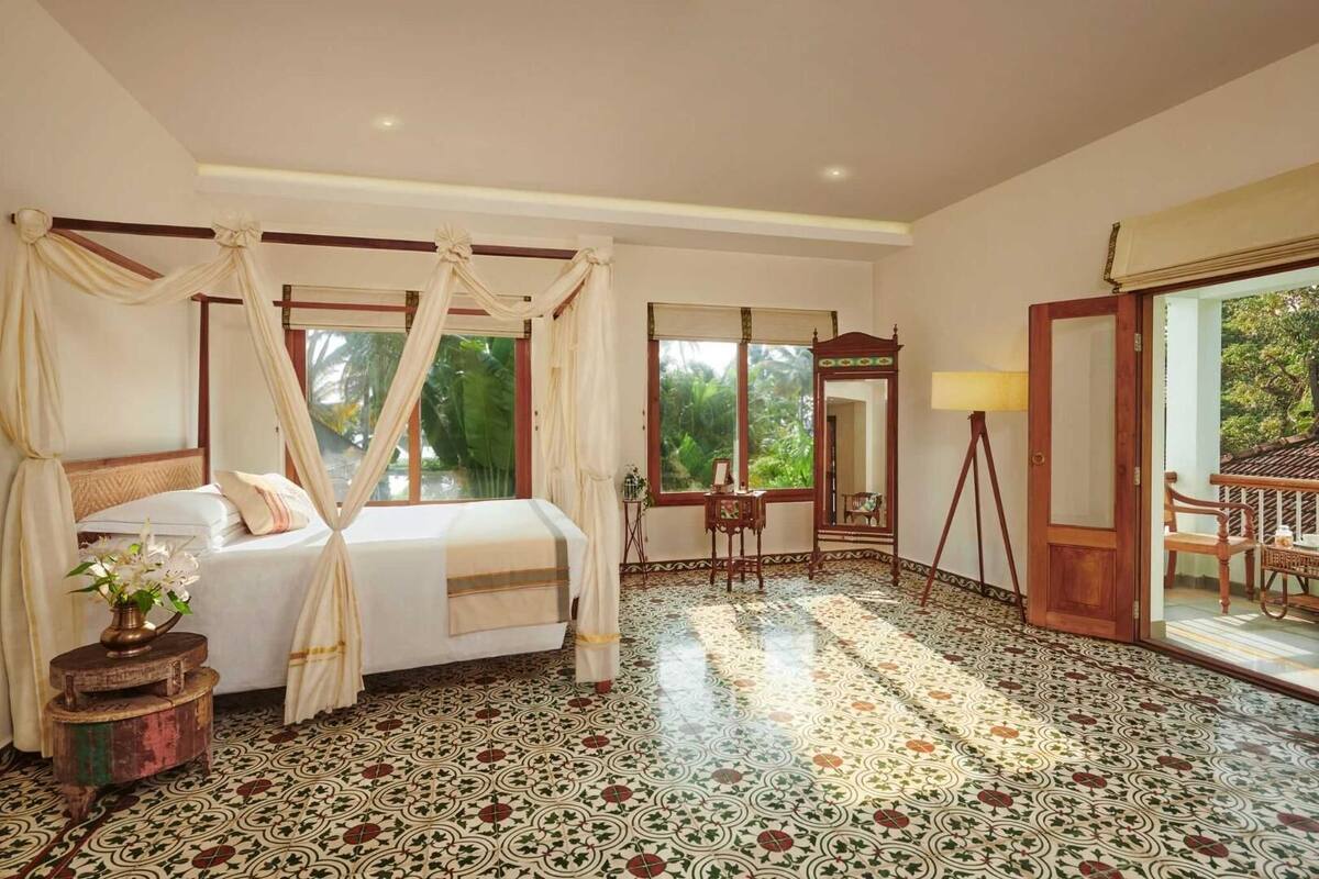 A bedroom with a bed, a bedside table, a mirror, a lamp, and a view of the outdoors.
