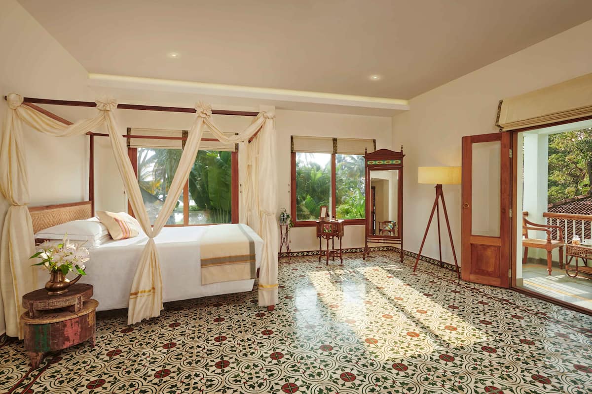 A spacious bedroom with a large bed, a small table with a lamp, and a view of the outdoors through a window.
