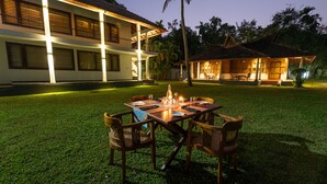 Uma área de refeições ao ar livre com uma mesa posta para dois, rodeada por cadeiras, em frente a um edifício com telhado em azulejos e um espaço coberto para sentar.