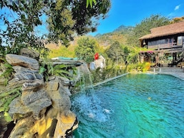 Piscina cristalina con cascada, rodeada de exuberante vegetación y montañas al fondo.