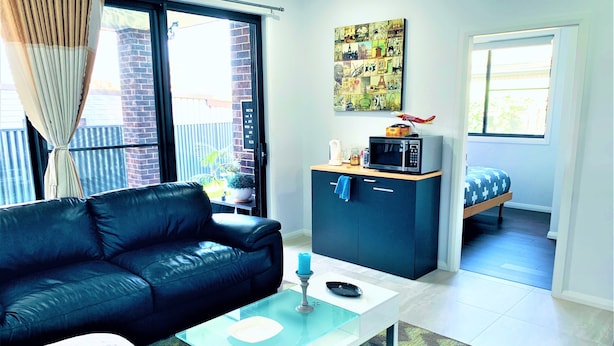 A compact living space with a black leather sofa, a glass coffee table, a kitchenette with a microwave, and a bed with patterned bedding.