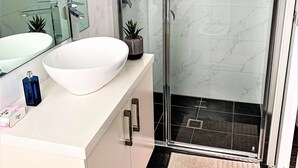 A modern bathroom with a large mirror, a white sink, a glass shower enclosure, and a small potted plant.