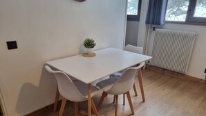 Een kleine, moderne kamer met een witte tafel en vier stoelen, een kleine potplant en een raam met gordijnen.