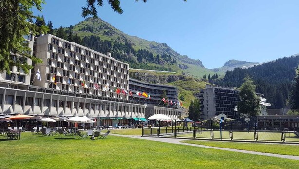 Een hotel met meerdere vlaggen, terras en een bergachtige achtergrond.