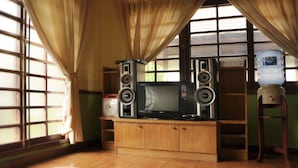 A room with a TV on a wooden cabinet, two speakers, a water dispenser, and large windows with curtains. - VILLA ADE - 0620 KOTA BUNGA PUNCAK (Cipanas)
